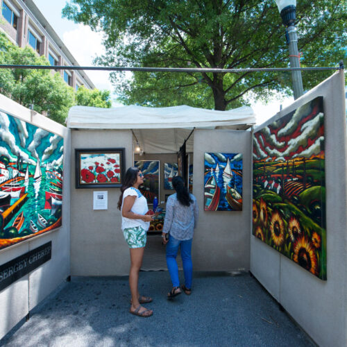 two women look at large paintings in art booth on street