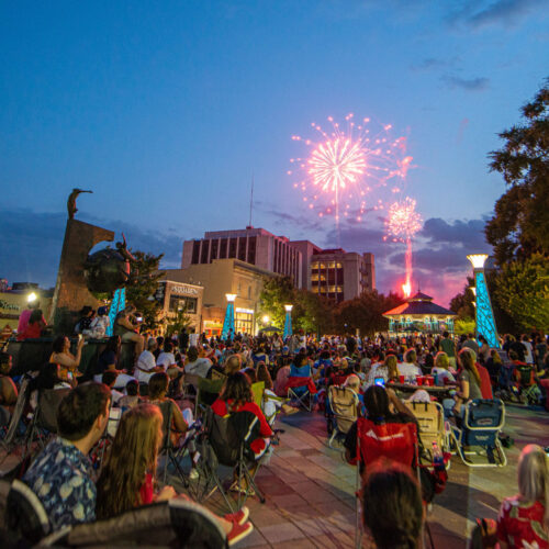 crowd filled downtown square with fireworks bursting overhead on July 4