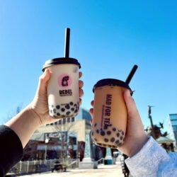 Two Hands Hold Up Rebel Teahouse Bubble Tea Drinks In Front Of Blue Sky