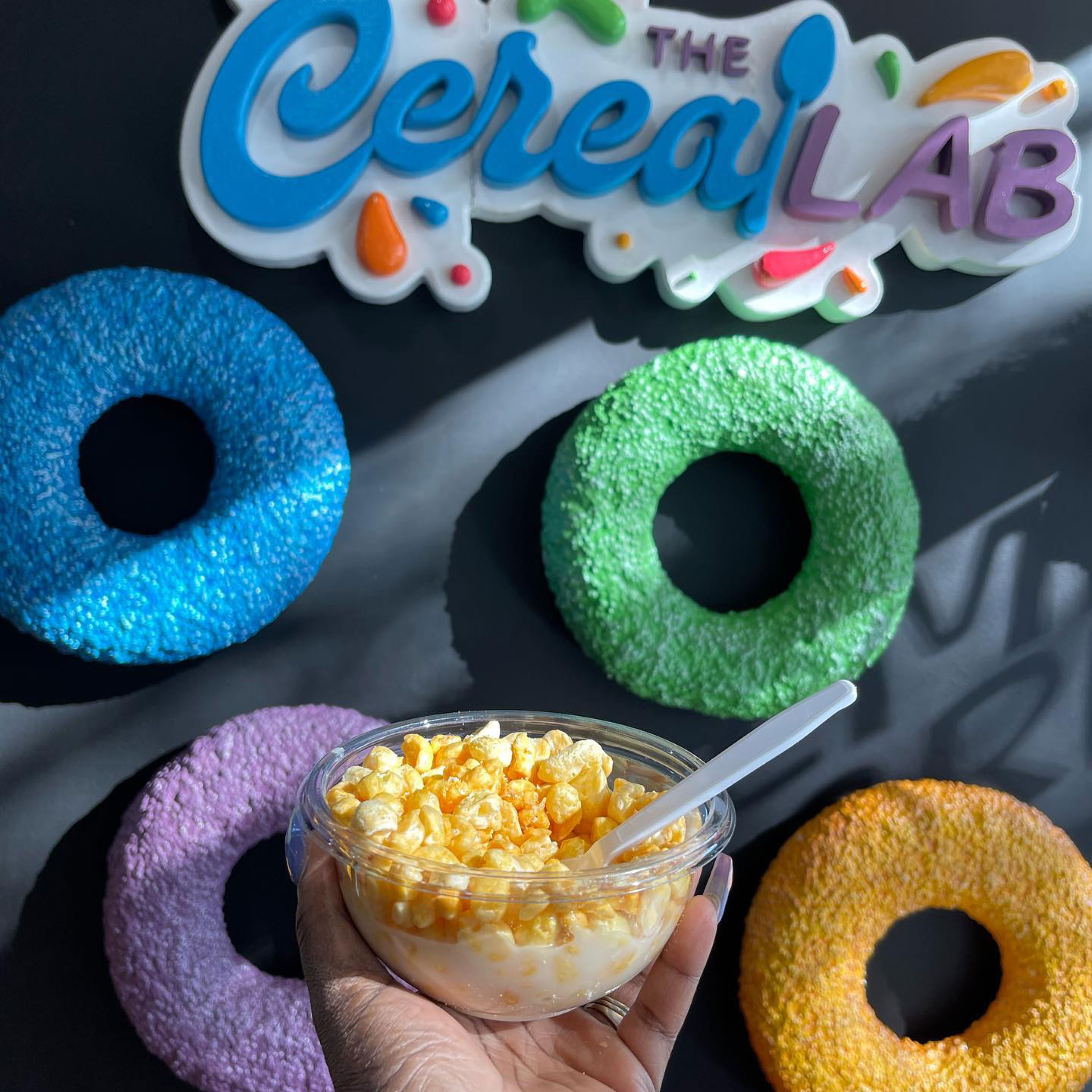 hand holding bowl of cereal in front of "Cereal Lab" sign and giant colorful fruit loops